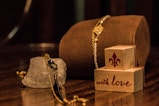 A composition of elegant jewelry placed on a reflective wooden surface. A gold and black bracelet drapes over a rough, translucent crystal. Nearby, two wooden blocks are stacked, featuring a fleur-de-lis symbol and the phrase 'with love' in decorative script.