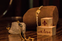 A composition of elegant jewelry placed on a reflective wooden surface. A gold and black bracelet drapes over a rough, translucent crystal. Nearby, two wooden blocks are stacked, featuring a fleur-de-lis symbol and the phrase 'with love' in decorative script.