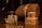 A composition of elegant jewelry placed on a reflective wooden surface. A gold and black bracelet drapes over a rough, translucent crystal. Nearby, two wooden blocks are stacked, featuring a fleur-de-lis symbol and the phrase 'with love' in decorative script.