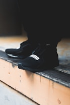 A close-up view of black athletic shoes with a visible brand logo, placed on a concrete step. The shoes are stylishly designed and appear to be suitable for sports or casual wear. The background is blurred, focusing attention on the footwear.
