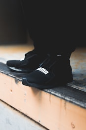 A close-up view of black athletic shoes with a visible brand logo, placed on a concrete step. The shoes are stylishly designed and appear to be suitable for sports or casual wear. The background is blurred, focusing attention on the footwear.