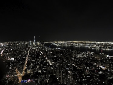 A nighttime cityscape featuring a densely packed skyline with a river running through the city. Numerous skyscrapers are lit up, and streets are illuminated with streetlights and car lights, creating a vibrant urban glow. The tallest building has a distinct spire, standing prominently among other high-rise structures.