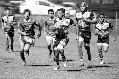 A group of rugby players in motion on a field, with one player prominently holding a rugby ball and running forward while others chase behind. The background features a blurred view of cars and a fence.