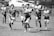 A group of rugby players in motion on a field, with one player prominently holding a rugby ball and running forward while others chase behind. The background features a blurred view of cars and a fence.