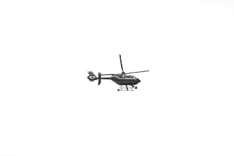A black helicopter with visible rotors and landing skids is flying against a stark white background. The helicopter is detailed and appears to have a word inscribed on its side.
