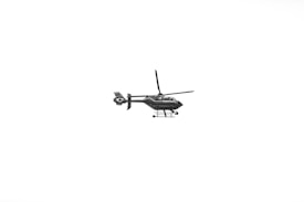 A black helicopter with visible rotors and landing skids is flying against a stark white background. The helicopter is detailed and appears to have a word inscribed on its side.