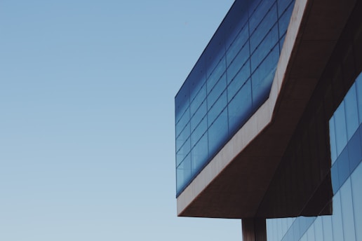 A modern building with large glass windows reflecting a clear blue sky. The structure features sharp angles and a minimalist design, emphasizing clean lines and a contemporary architectural style.