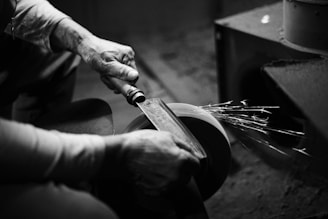 A person is sharpening a blade on a grinding wheel, with sparks flying off from the friction. The hands and tool are in focus, while the surroundings are blurred, giving an intimate, detailed view of the activity.