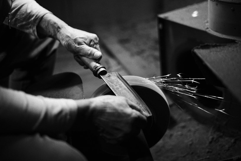 A person is sharpening a blade on a grinding wheel, with sparks flying off from the friction. The hands and tool are in focus, while the surroundings are blurred, giving an intimate, detailed view of the activity.