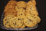 A clear glass tray holds an assortment of freshly baked cookies, including oatmeal raisin, chocolate chip, and nutty varieties, each featuring a golden-brown hue. The cookies are neatly arranged in rows on the tray against a dark backdrop, highlighting their textures.