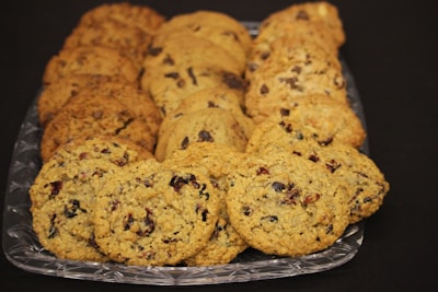 A clear glass tray holds an assortment of freshly baked cookies, including oatmeal raisin, chocolate chip, and nutty varieties, each featuring a golden-brown hue. The cookies are neatly arranged in rows on the tray against a dark backdrop, highlighting their textures.