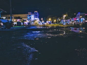 A nighttime cityscape with wet pavement reflecting artificial lights from buildings and street lamps. The scene includes parked cars and illuminated storefronts, creating a vibrant urban atmosphere.