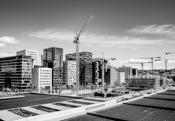 A cityscape with a collection of modern buildings featuring various architectural designs. Tower cranes are visible, suggesting ongoing construction projects. The scene is captured in black and white, providing a stark yet elegant view of urban development.