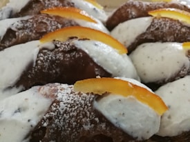A close-up view of multiple pastries topped with white cream filling and garnished with slices of candied orange peel. The pastries have a dusting of powdered sugar, and the texture of the pastry is flaky and golden brown.