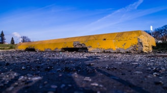 A close-up view of a damaged yellow concrete parking curb lying on a rough, gravelly surface. The curb shows significant wear and erosion, with chunks missing and a rough texture. The background consists of a blue sky with wispy clouds and some sparse trees.