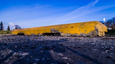 A close-up view of a damaged yellow concrete parking curb lying on a rough, gravelly surface. The curb shows significant wear and erosion, with chunks missing and a rough texture. The background consists of a blue sky with wispy clouds and some sparse trees.