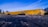 A close-up view of a damaged yellow concrete parking curb lying on a rough, gravelly surface. The curb shows significant wear and erosion, with chunks missing and a rough texture. The background consists of a blue sky with wispy clouds and some sparse trees.