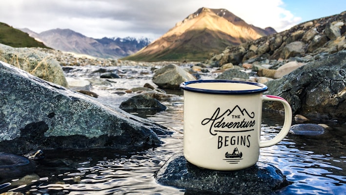 A ceramic mug with a motivational message rests on a rock in a shallow stream. The riverbed is filled with smooth stones, and the background features a serene mountain landscape under a partly cloudy sky.