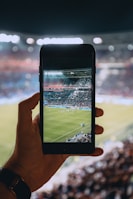 A hand holding a smartphone captures a live view of a soccer stadium filled with fans and players on the field. The image on the smartphone screen is crisp, showing a wide-angle view of the game in action.