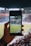 A hand holding a smartphone captures a live view of a soccer stadium filled with fans and players on the field. The image on the smartphone screen is crisp, showing a wide-angle view of the game in action.