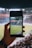 A hand holding a smartphone captures a live view of a soccer stadium filled with fans and players on the field. The image on the smartphone screen is crisp, showing a wide-angle view of the game in action.