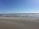 A sandy beach stretches into the distance with gentle ocean waves in the background. Several kite surfers are visible with colorful kites flying in the sky under a bright blue atmosphere.