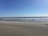 A sandy beach stretches into the distance with gentle ocean waves in the background. Several kite surfers are visible with colorful kites flying in the sky under a bright blue atmosphere.