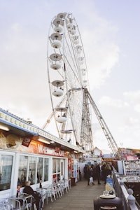 A large Ferris wheel towers over a bustling boardwalk area. People are gathered around, some seated outside a cafe with a bright red and white sign that reads 'Whemoo's Cafe.' The atmosphere is lively with a variety of colorful shops and attractions nearby.