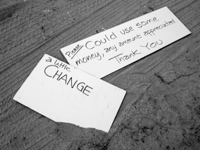 Two cardboard signs rest on the ground. One sign reads 'a little CHANGE' in black lettering, while the other says 'Please Could use some money, any amount appreciated. Thank You.' The background is textured sand.