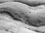 A monochrome image with a textured rocky surface. The landscape is composed of smooth, undulating formations with scattered small holes and pebbles. It appears to be a natural terrain with a lunar or desert-like quality.