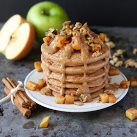A stack of pancakes topped with a generous amount of almond or peanut butter sauce, garnished with chunks of caramelized apples and nuts. A fresh green apple is visible in the background alongside a sliced half of an apple. Cinnamon sticks are tied together with a string nearby, enhancing the rustic and cozy presentation.