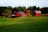 A vibrant red barn and adjacent building are situated on a serene, green hillside, surrounded by lush trees. The foreground features open, grassy land dotted with a few grazing animals. An autumn tree with orange leaves contrasts with the green landscape, and a farmhouse is visible in the distance.