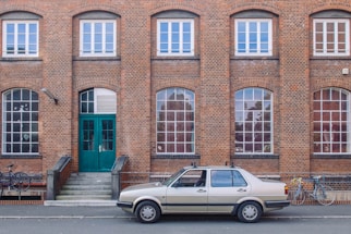 A vintage beige car is parked in front of a brick building with several large, arched windows. The building features a central green double door accessed by a short set of steps. Two bicycles are visible to the side of the car.