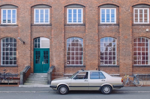 A vintage beige car is parked in front of a brick building with several large, arched windows. The building features a central green double door accessed by a short set of steps. Two bicycles are visible to the side of the car.