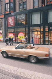 A blurred brown convertible car is driving past a modern building with large glass windows. The building has prominent sale signs displaying a 50% discount. Several pedestrians are walking on the sidewalk.