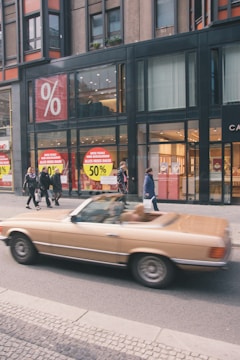 A blurred brown convertible car is driving past a modern building with large glass windows. The building has prominent sale signs displaying a 50% discount. Several pedestrians are walking on the sidewalk.