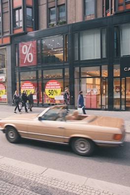 A blurred brown convertible car is driving past a modern building with large glass windows. The building has prominent sale signs displaying a 50% discount. Several pedestrians are walking on the sidewalk.