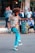 A man with curly hair, wearing a colorful t-shirt featuring various faces and blue jeans, is performing a street act. He is holding sticks and appears to be controlling a yo-yo or similar item. Several people are observing, some standing while others sit in the background. One observer is taking a picture with a phone, and there is a metal trash can nearby.