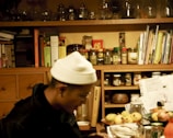A cozy kitchen space with wooden shelves filled with various jars and containers. Cookbooks and papers are scattered on the shelves. A person sitting at a table with a white hat on is slightly blurred, surrounded by fruits and bottles.