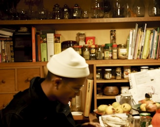A cozy kitchen space with wooden shelves filled with various jars and containers. Cookbooks and papers are scattered on the shelves. A person sitting at a table with a white hat on is slightly blurred, surrounded by fruits and bottles.