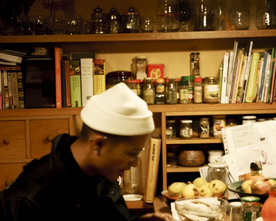 A cozy kitchen space with wooden shelves filled with various jars and containers. Cookbooks and papers are scattered on the shelves. A person sitting at a table with a white hat on is slightly blurred, surrounded by fruits and bottles.