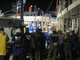 A group of people, including some wearing uniforms labeled 'MARINA,' are gathered around a partially collapsed structure at night. Many are wearing safety helmets and other protective gear, suggesting an emergency response or rescue operation. The scene is illuminated by artificial lighting, and some individuals are on top of the debris while others are standing nearby.