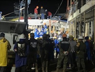 A group of people, including some wearing uniforms labeled 'MARINA,' are gathered around a partially collapsed structure at night. Many are wearing safety helmets and other protective gear, suggesting an emergency response or rescue operation. The scene is illuminated by artificial lighting, and some individuals are on top of the debris while others are standing nearby.