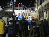 A group of people, including some wearing uniforms labeled 'MARINA,' are gathered around a partially collapsed structure at night. Many are wearing safety helmets and other protective gear, suggesting an emergency response or rescue operation. The scene is illuminated by artificial lighting, and some individuals are on top of the debris while others are standing nearby.