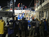 A group of people, including some wearing uniforms labeled 'MARINA,' are gathered around a partially collapsed structure at night. Many are wearing safety helmets and other protective gear, suggesting an emergency response or rescue operation. The scene is illuminated by artificial lighting, and some individuals are on top of the debris while others are standing nearby.