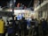 A group of people, including some wearing uniforms labeled 'MARINA,' are gathered around a partially collapsed structure at night. Many are wearing safety helmets and other protective gear, suggesting an emergency response or rescue operation. The scene is illuminated by artificial lighting, and some individuals are on top of the debris while others are standing nearby.