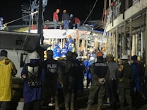 A group of people, including some wearing uniforms labeled 'MARINA,' are gathered around a partially collapsed structure at night. Many are wearing safety helmets and other protective gear, suggesting an emergency response or rescue operation. The scene is illuminated by artificial lighting, and some individuals are on top of the debris while others are standing nearby.