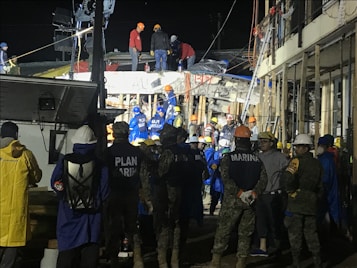 A group of people, including some wearing uniforms labeled 'MARINA,' are gathered around a partially collapsed structure at night. Many are wearing safety helmets and other protective gear, suggesting an emergency response or rescue operation. The scene is illuminated by artificial lighting, and some individuals are on top of the debris while others are standing nearby.