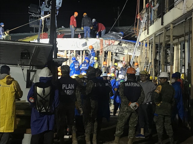 A group of people, including some wearing uniforms labeled 'MARINA,' are gathered around a partially collapsed structure at night. Many are wearing safety helmets and other protective gear, suggesting an emergency response or rescue operation. The scene is illuminated by artificial lighting, and some individuals are on top of the debris while others are standing nearby.