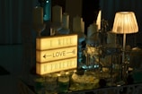 A warmly lit scene features a light box displaying the text 'I will love you forever' with an arrow pointing to the word 'love.' Surrounding the light box are several elegant white candles, an intricately designed birdcage, decorative glass bowls with flowers floating in them, and a beige lamp. The arrangement suggests a romantic or intimate setting.